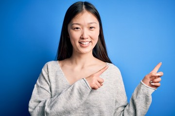 Sticker - Young beautiful asian woman wearing casual sweater standing over blue isolated background smiling and looking at the camera pointing with two hands and fingers to the side.