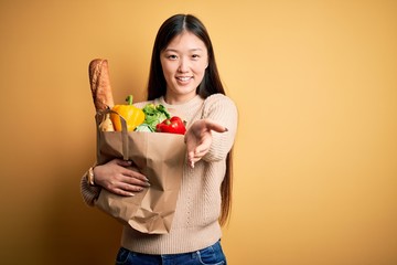 Sticker - Young asian woman holding paper bag of fresh healthy groceries over yellow isolated background smiling cheerful offering palm hand giving assistance and acceptance.