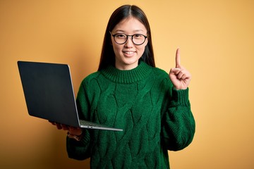 Young asian business woman wearing glasses and working using computer laptop with a big smile on face, pointing with hand finger to the side looking at the camera.