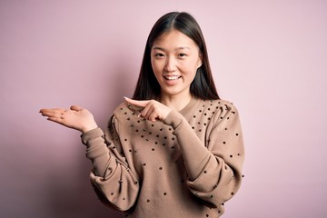 Sticker - Young beautiful asian woman wearing fashion and elegant sweater over pink solated background amazed and smiling to the camera while presenting with hand and pointing with finger.