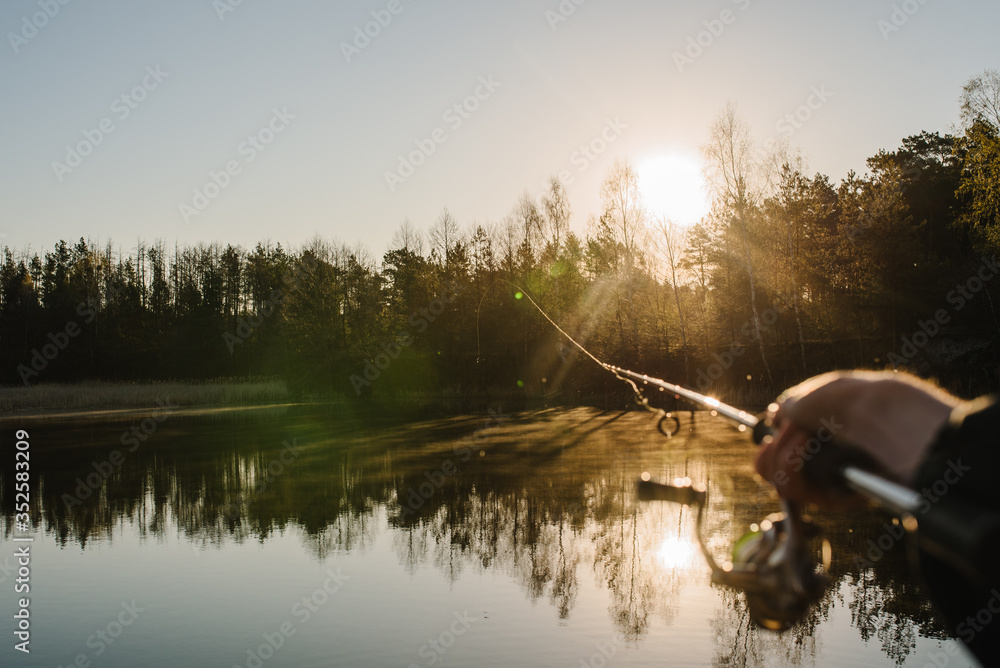 Fisherman with rod throws bait into the water on river bank. Fishing for pike, perch. Background wild nature. Man catching a fish on lake or pond with text space. Fishing day. Misty morning. Sunrise. - obrazy, fototapety, plakaty 