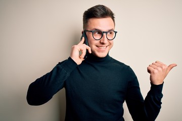 Wall Mural - Young handsome caucasian man having a conversation talking on smartphone pointing and showing with thumb up to the side with happy face smiling