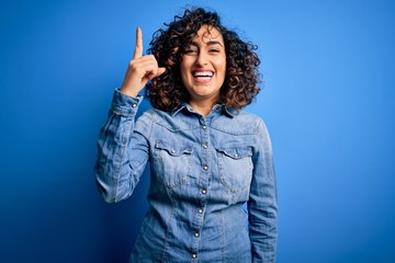 Poster - Young beautiful curly arab woman wearing casual denim shirt standing over blue background pointing finger up with successful idea. Exited and happy. Number one.