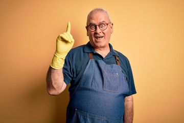 Wall Mural - Middle age cleaner man cleaning wearing apron and gloves over isolated yellow background pointing finger up with successful idea. Exited and happy. Number one.