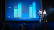 © Gorodenkoff - On Stage, Successful Female Executive and Male Manager Present New Product, Show Infographics, Statistics on Big Screen, Talk About Growth. Live Event, Tech Startup, Business Conference