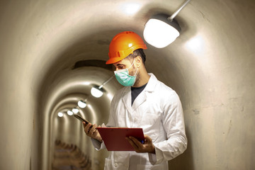 Tecnico con caschetto protettivo e camice bianco guarda un messaggio dal suo cellulare dentro un tunnel sotterraneo