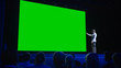 © Gorodenkoff - Keynote Speaker Does Presentation of New Product to the Audience, Behind Him Movie Theater with Green Screen, Mock-up, Chroma Key. Business Conference Live Event or Device Reveal