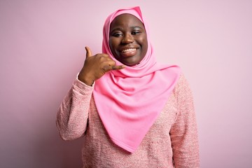 Poster - Young african american plus size woman wearing muslim hijab over isolated pink background smiling doing phone gesture with hand and fingers like talking on the telephone. Communicating concepts.