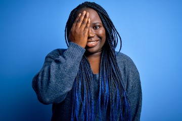 Sticker - African american plus size woman with braids wearing casual sweater over blue background covering one eye with hand, confident smile on face and surprise emotion.