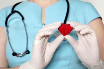 Woman doctor in medical mask holding red knitted heart in hands, saving lives during a covid-19 coronavirus pandemic. Concept of cardiologist, blood donation, treatment in clinic