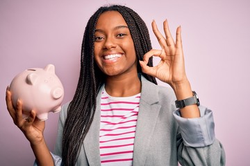 Poster - Young african american business woman saving money on piggy bank over isolated background doing ok sign with fingers, excellent symbol