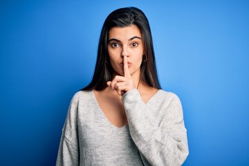 Wall Mural - Young beautiful brunette woman wearing casual sweater standing over blue background asking to be quiet with finger on lips. Silence and secret concept.