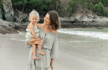 Cute Happy Mom Model And Her Blonde Daughter In Her Arms Walk Along A Tropical Beach In Summer Enjoying A Vacation.