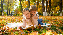 Adorable Toddler Boy Sitting On Mothers Back And Reading A Book At Autumn Park