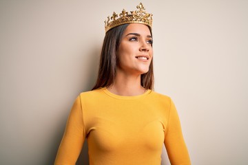 Wall Mural - Young beautiful brunette woman wearing golden queen crown over white background looking away to side with smile on face, natural expression. Laughing confident.