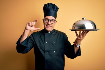 Sticker - Middle age handsome grey-haired waiter man wearing cooker uniform and hat holding tray smiling and confident gesturing with hand doing small size sign with fingers looking and the camera. Measure