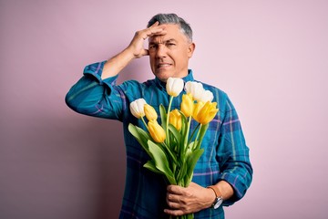 Wall Mural - Middle age grey-haired man holding natural bouquet of yellow tulips over pink background stressed with hand on head, shocked with shame and surprise face, angry and frustrated. Fear and upset