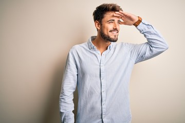 Wall Mural - Young handsome man with beard wearing striped shirt standing over white background very happy and smiling looking far away with hand over head. Searching concept.