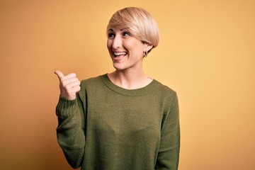 Poster - Young blonde woman with modern short hair wearing casual sweater over yellow background smiling with happy face looking and pointing to the side with thumb up.