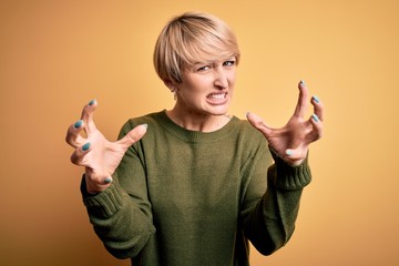 Wall Mural - Young blonde woman with modern short hair wearing casual sweater over yellow background Shouting frustrated with rage, hands trying to strangle, yelling mad