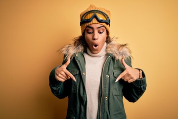 Wall Mural - Young african american afro skier girl wearing snow sportswear and ski goggles Pointing down with fingers showing advertisement, surprised face and open mouth