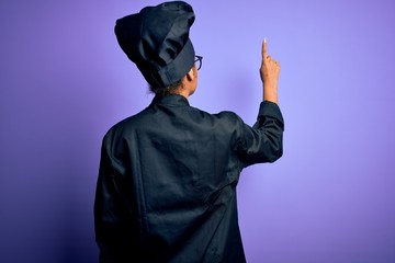 Sticker - Young african american chef girl wearing cooker uniform and hat over purple background Posing backwards pointing ahead with finger hand