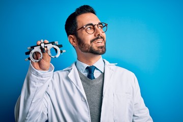 Poster - Young handsome optical man with beard holding optometry glasses over blue background looking away to side with smile on face, natural expression. Laughing confident.