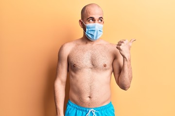 Sticker - Young handsome bald man shirtless wearing swimwear and medical mask pointing thumb up to the side smiling happy with open mouth