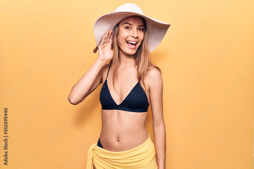 Young beautiful girl wearing bikini and hat smiling with hand over ear listening and hearing to rumor or gossip. deafness concept. - obrazy, fototapety, plakaty 