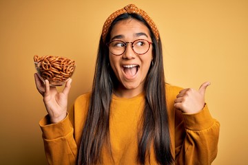 Canvas Print - Young beautiful asian woman holding bowl with german baked pretzels over yellow background pointing and showing with thumb up to the side with happy face smiling