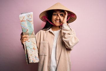 Canvas Print - Young thai tourist woman on vacation wearing traditional conical asian hat holding city map with happy face smiling doing ok sign with hand on eye looking through fingers