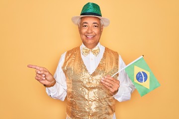 Poster - Middle age senior grey-haired man wearing Brazilian carnival custome holding Brazil flag very happy pointing with hand and finger to the side