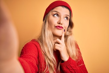 Poster - Young beautiful blonde woman making selfie by camera over isolated yellow background serious face thinking about question, very confused idea