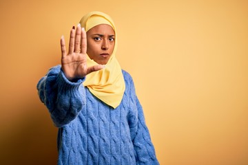 Wall Mural - Young African American afro woman wearing muslim hijab over isolated yellow background doing stop sing with palm of the hand. Warning expression with negative and serious gesture on the face.