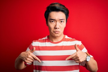 Wall Mural - Young handsome chinese man wearing casual striped t-shirt standing over red background Pointing down looking sad and upset, indicating direction with fingers, unhappy and depressed.