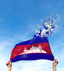 Two hands are holding a Cambodia flag which morphs into birds while waving against a blue sky background. 3D Illustration.