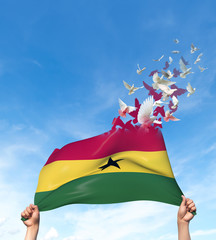 Two hands are holding a Ghana flag which morphs into birds while waving against a blue sky background. 3D Illustration.