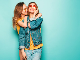 Two young beautiful smiling hipster girls in trendy summer yellow T-shirts and jeans jacket. Sexy carefree women posing near blue wall. Trendy and positive models having fun