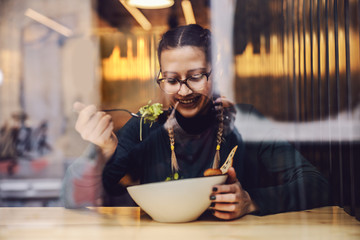 Young smiling girl sitting in restaurant and enjoying her salad for lunch. healthy lifestyle concept. Picture taken from outside.