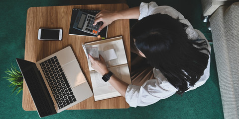 Above shot woman calculating and holding an Invoice while sitting at the wooden short legs table that surrounded by laptop and equipment over comfortable sitting room as background.