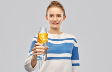 Wall Mural - drink, health and people concept - happy smiling teenage girl in sweater with orange fruit infused water in glass bottle over grey background