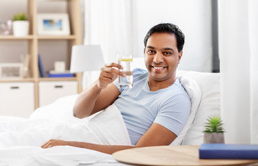 Poster - people, bedtime and rest concept - happy smiling indian man lying in bed at home and taking glass of water with lemon from table