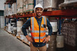 © StratfordProductions - Portrait of happy manager standing with his hands on waist wearing white helmet and safety vest in warehouse with shelves of inventory in the background