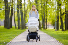 Young Mother Pushing White Baby Stroller And Slowly Walking At Town Green Park In Warm, Sunny Summer Day. Spending Time With Infant And Breathing Fresh Air. Enjoying Stroll. Front View.
