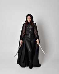 Wall Mural - fantasy portrait of a woman with red hair wearing dark leather assassin costume with long black cloak. Full length standing pose holding a sword isolated against a studio background.