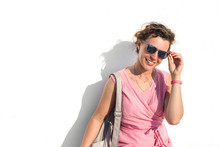 Portrait Attractive Woman In Red White Striped Dress,wearing Sunglasses,backpack Sitting Near White Wall Background In Summer Sunny Day. Caucasian Curly Female Student Looking At Camera Smiling