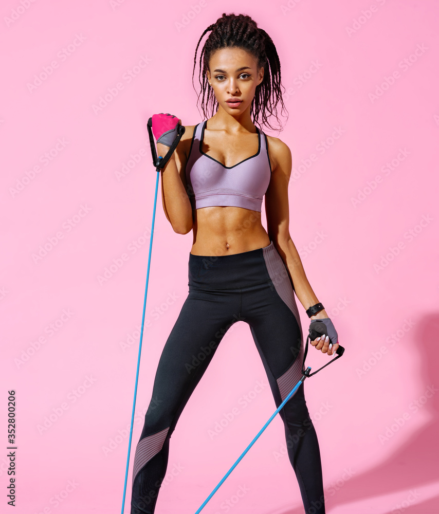 Strong girl working with resistance band. Photo of african american girl in fashionable sportswear on pink background. Sports and healthy lifestyle - obrazy, fototapety, plakaty 
