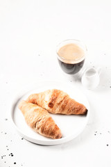 Two croissants on a plate next to a cup of coffee on light gray background.