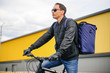 © ART_ist - Courier delivery service. A man in glasses and a windbreaker rides a bicycle with a backpack along a city street against the background of the building. Online food and goods orders