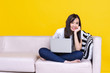 © jumlongch - Asian woman smiling and working from home use laptop computer while sitting on sofa over isolate yellow background